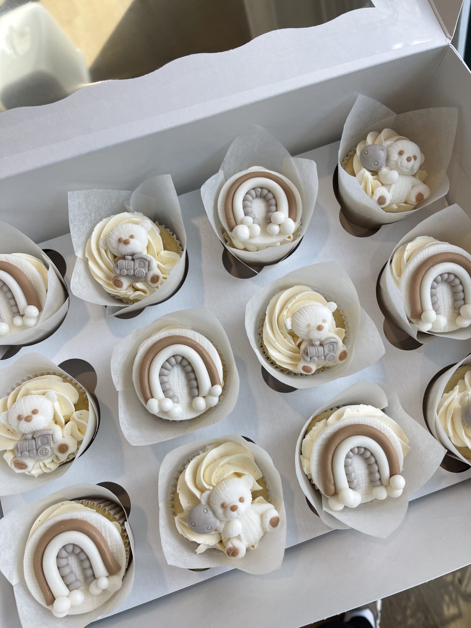 Gender neutral coloured cupcakes topped with fondant rainbows, teddy bears, and baby‑themed decorations.