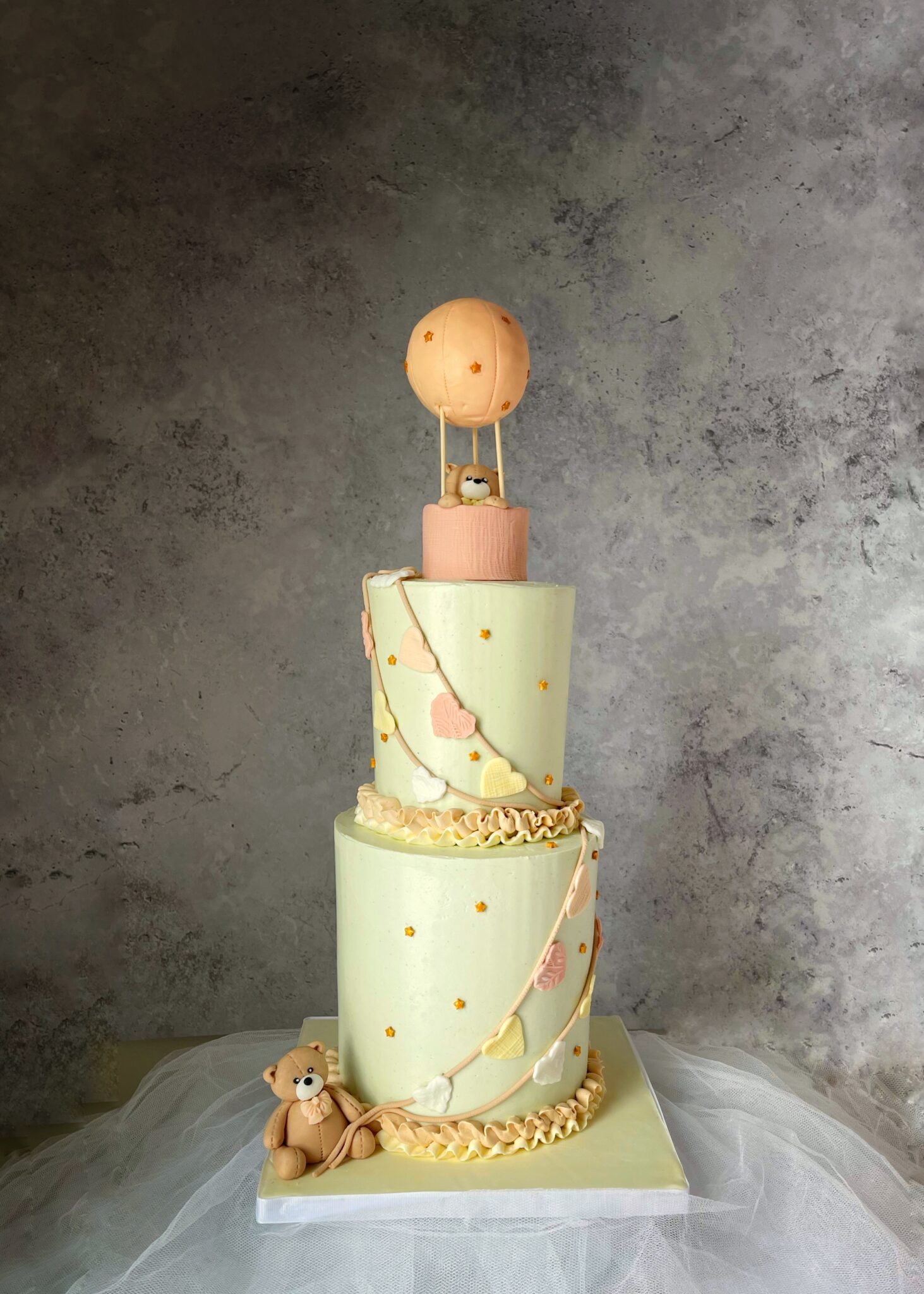 A three‑tier pastel cake decorated with clouds, stars, a teddy bear, and a small hot‑air‑balloon topper.