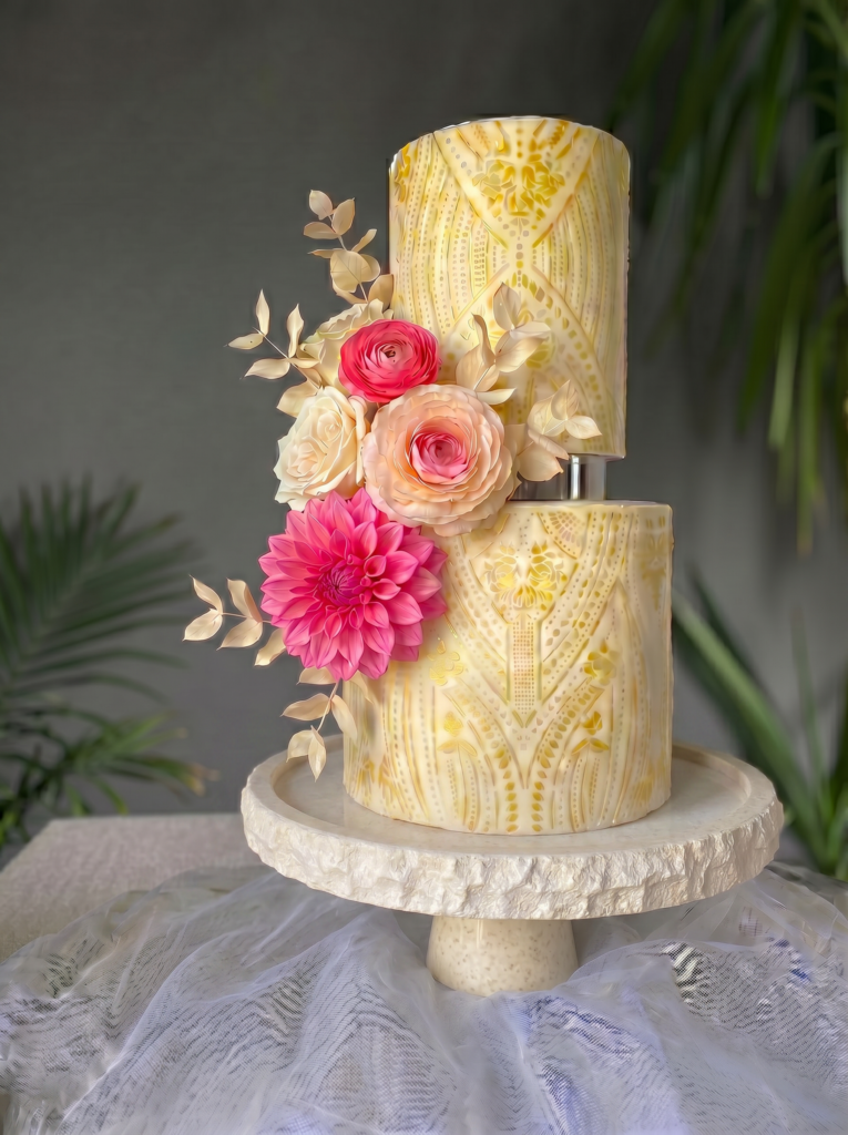 Two-tier ivory wedding cake with gold patterned detailing, blush and pink sugar flowers, displayed on a white pedestal.