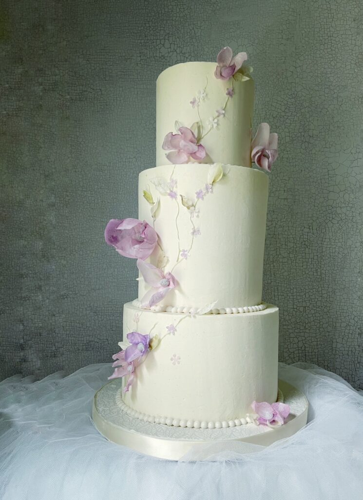 Elegant three-tier buttercream wedding cake with handmade blush and lavender wafer paper flowers cascading over smooth white tiers.
