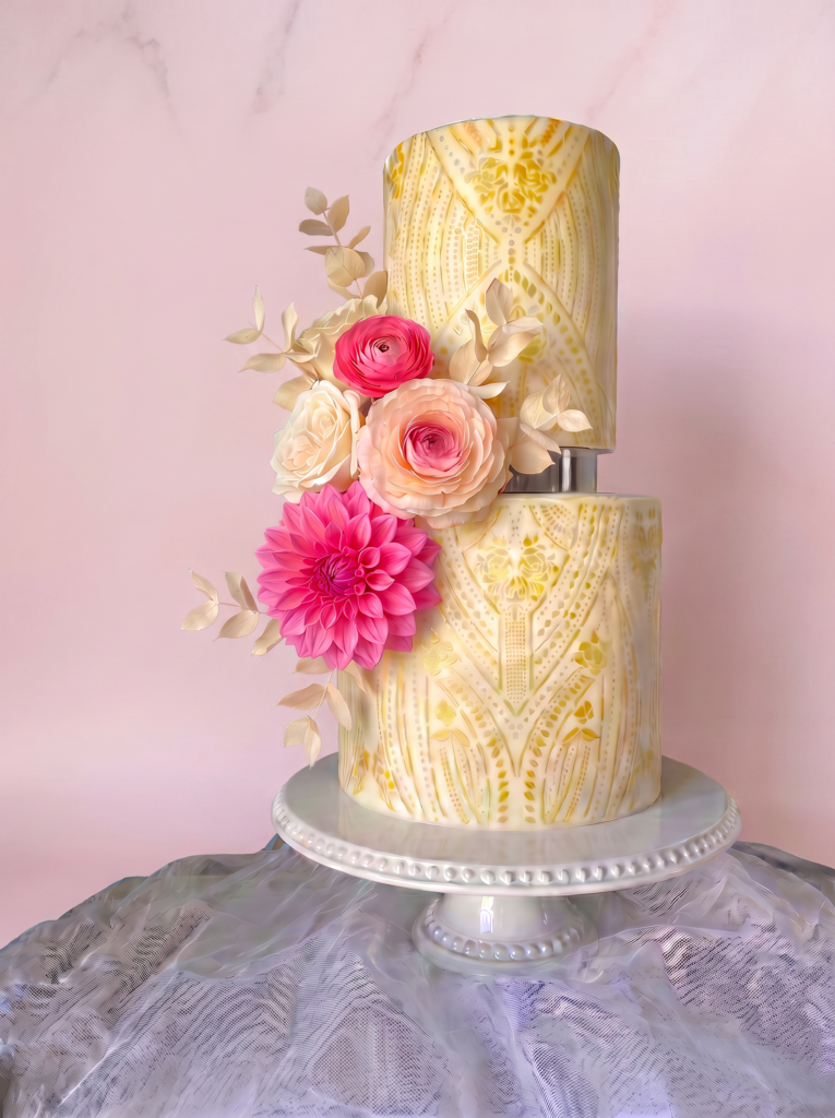 Two-tier ivory wedding cake with gold patterned detailing, blush and pink sugar flowers, displayed on a white pedestal.