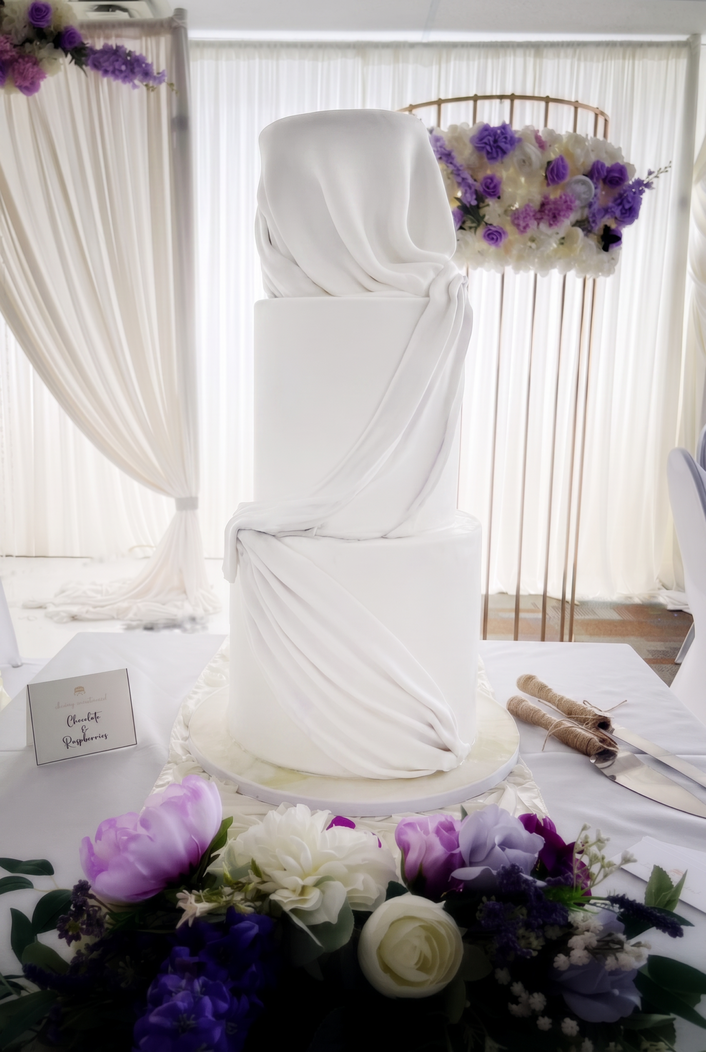 Three-tier white wedding cake featuring elegant draped fondant, surrounded by purple and white floral arrangements in a sophisticated wedding reception setting